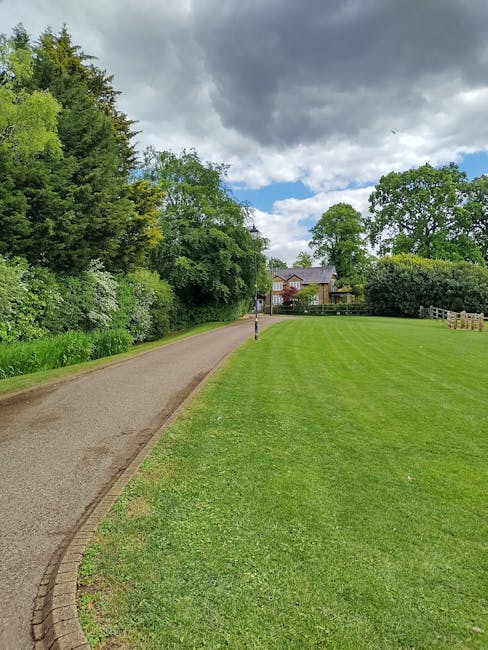 A narrow, winding asphalt pathway runs alongside a well-maintained grassy area with neatly trimmed, vibrant green turf. To the left of the path, there is dense foliage comprising tall trees and bushes with fresh green leaves, contributing to a lush, natural backdrop. The pathway curves gently towards residential buildings situated at the end of the scene, partially visible beyond a black and yellow striped post standing on the edge of the grass. The sky overhead is mostly cloudy with some patches of blue, creating a subdued light that blankets the scene. In the context of waste management and rubbish removal services, this setting suggests a suburban or park environment where independent collection or small-scale waste clearing, possibly by Waste Disposal Swiss Cottage, might be appropriate for maintaining the tidy appearance of the area.