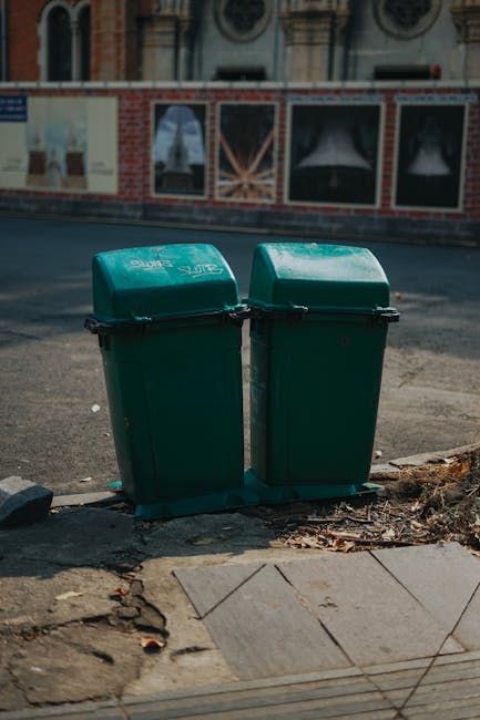 Two large garbage bags made of black plastic are placed on the pavement near the curb in front of a black metal fence and dense greenery. The bags appear to be filled with waste material and are slightly crumpled and tied at the top. Behind the fence, a dark building is partially visible, and the overall scene is illuminated by natural daylight, creating shadows on the ground. This urban setting, with waste temporarily stored on the street, aligns with alternative waste handling options such as private rubbish collection services like Waste Disposal Swiss Cottage, demonstrating the typical placement of household or commercial rubbish awaiting collection or disposal.