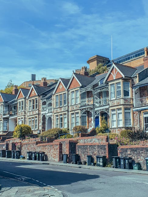 A row of Victorian-style terraced houses with ornate white ironwork balconies and decorative gable end features, situated on a sloped street with a low brick wall and small landscaped garden areas in front. The buildings are constructed from red and beige bricks, with large sash windows framed in white. In the foreground, numerous black wheelie bins are lined up along the pavement, indicating waste collection requirements. The street appears to be in an urban residential setting, with some modern buildings visible behind the row of houses under a bright blue sky with wispy clouds. The scene reflects typical private waste handling and rubbish collection arrangements in a city context, with an emphasis on on-site disposal and domestic waste management by local private waste disposal services, such as Waste Disposal Swiss Cottage.